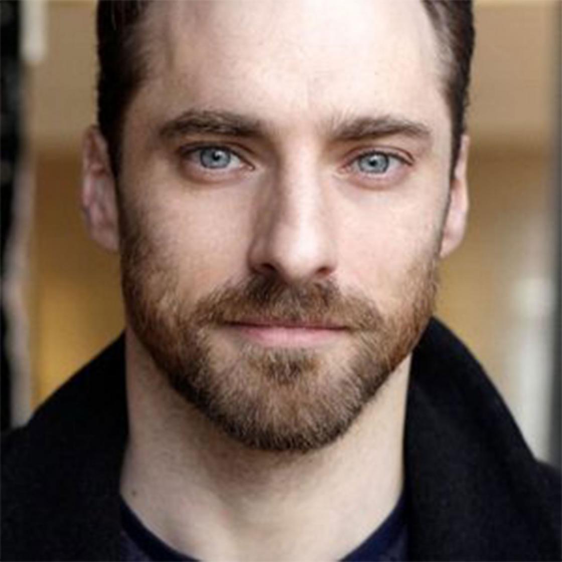 Richard Booth is a white male with dark brown hair and beard, he wears a black top