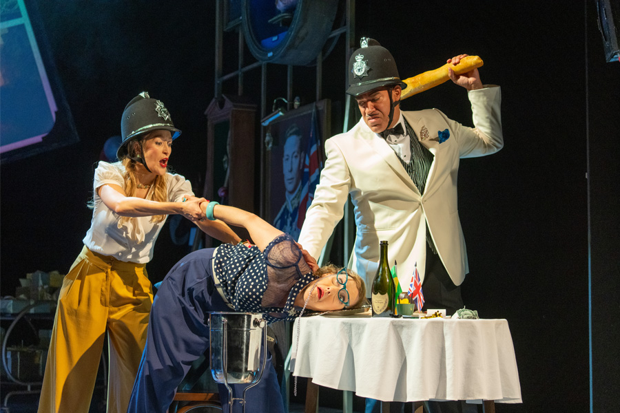 Two people wearing police hats push another against a table, threatening them with a baguette