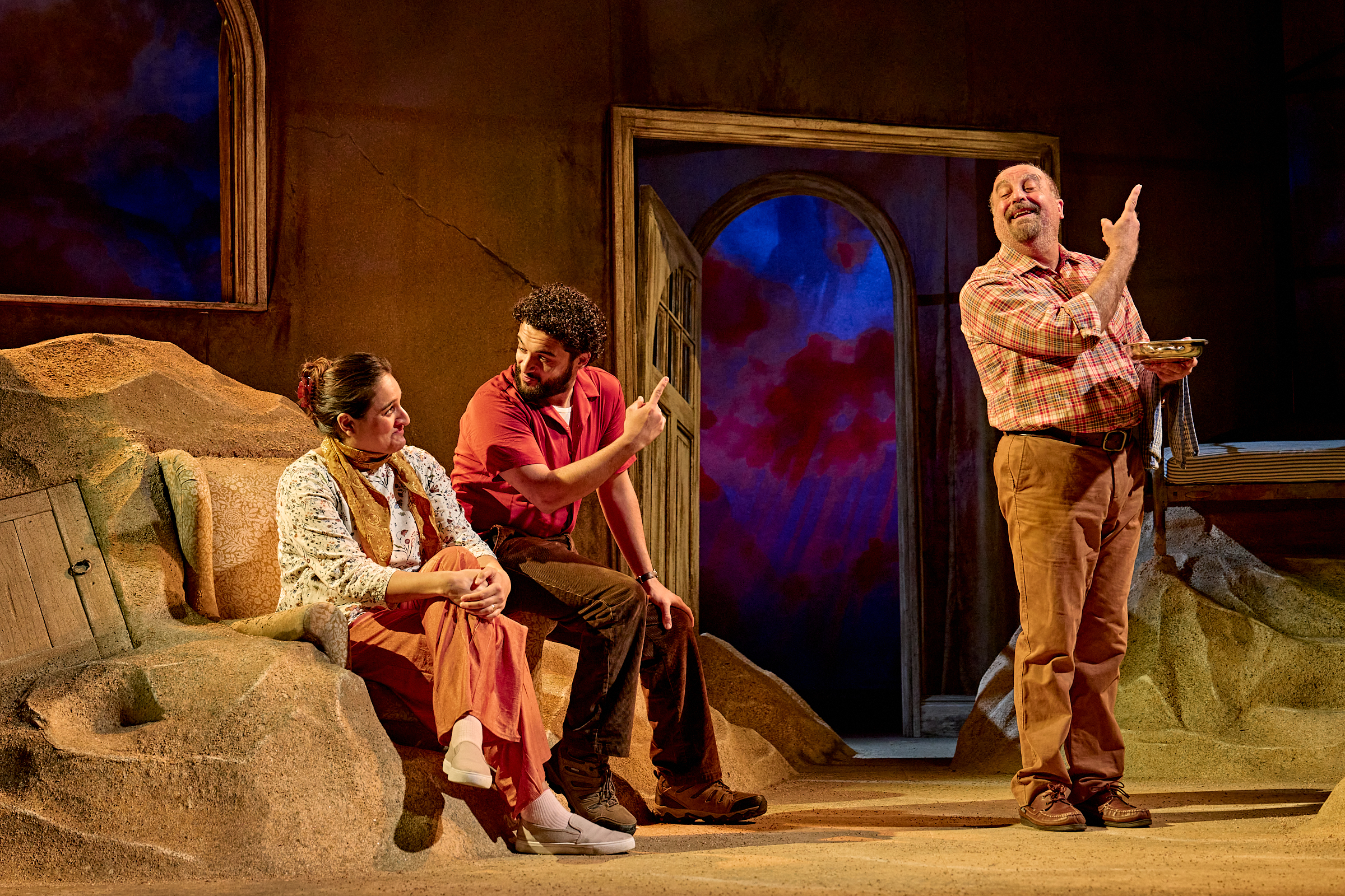 A man stands by a couple mid-speech, finger pointing to the sky, the couple are sitting. They look happy