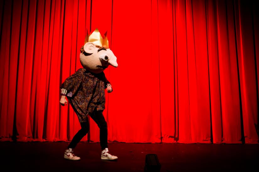 A man with a giant false head walks in front of a red curtain.