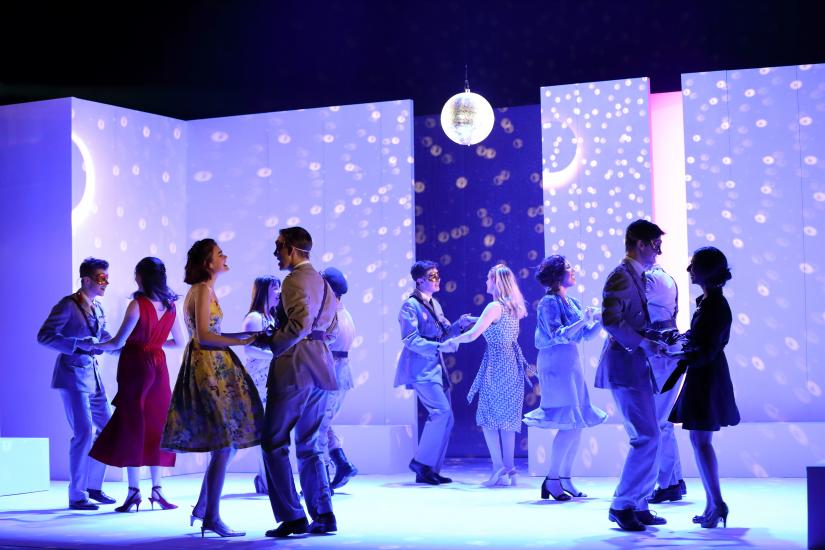 The cast ensemble dance in purple lighting with a glitterball
