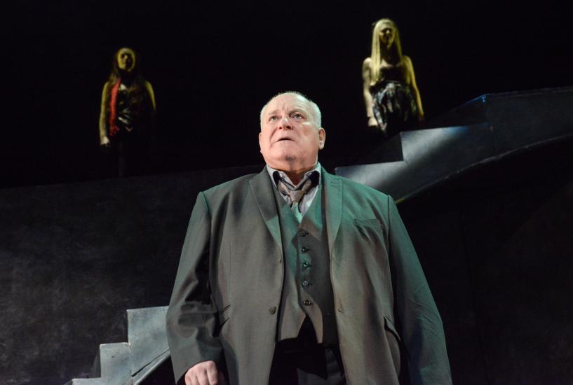 A man looks into the distance, two women stand on stairs behind him