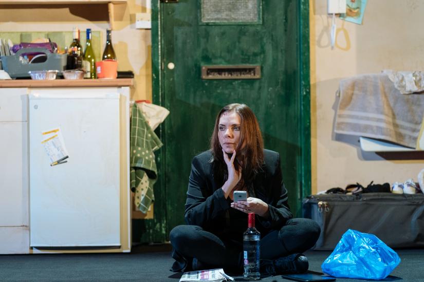 A woman sits cross-legged on the floor with a mobile phone in her hand, a bottle of vodka is next to her