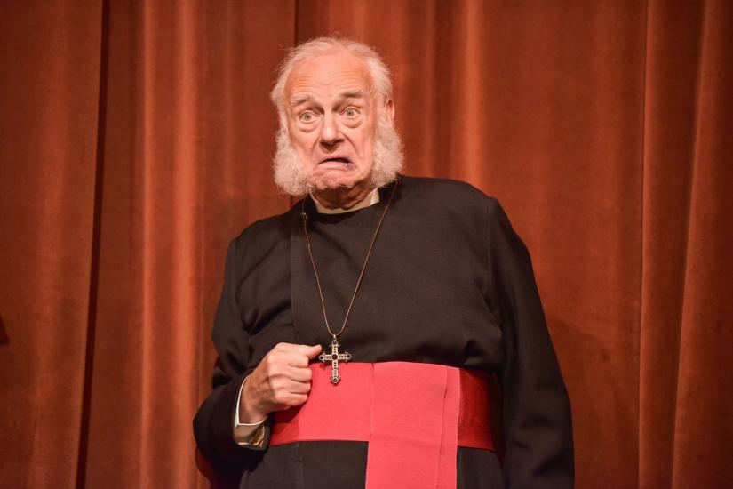 an old man in clergy dress with a red sash looks confused