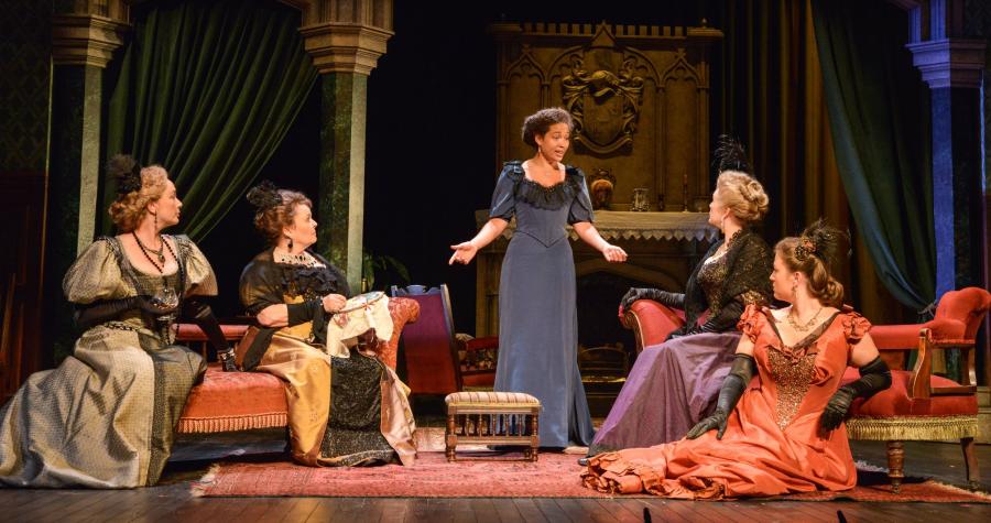 a group of women sit in a grand drawing room and listen to another woman