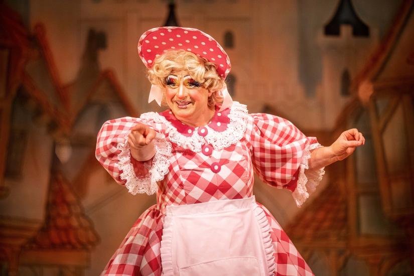 Dame Trott wears a pink gingham dress and matching hat, pointing to the audience