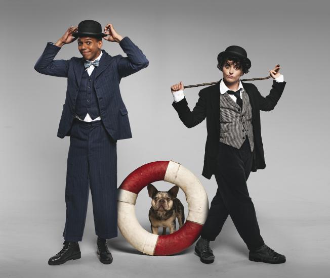 Charlie and Stan stand, one of them is holding a bowler hat on their head and the other is holding a cane behind their back. A French Bulldog is sitting on the floor and looking through a lifebuoy. 