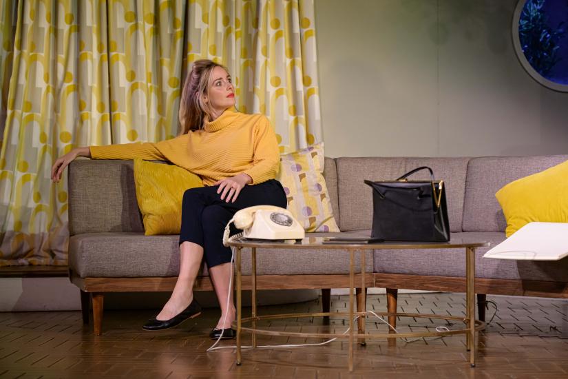 A woman sits on a sofa, with a telephone in front of her on a coffee table. She is wearing a mustard yellow jumper and black trousers, and she looks shocked