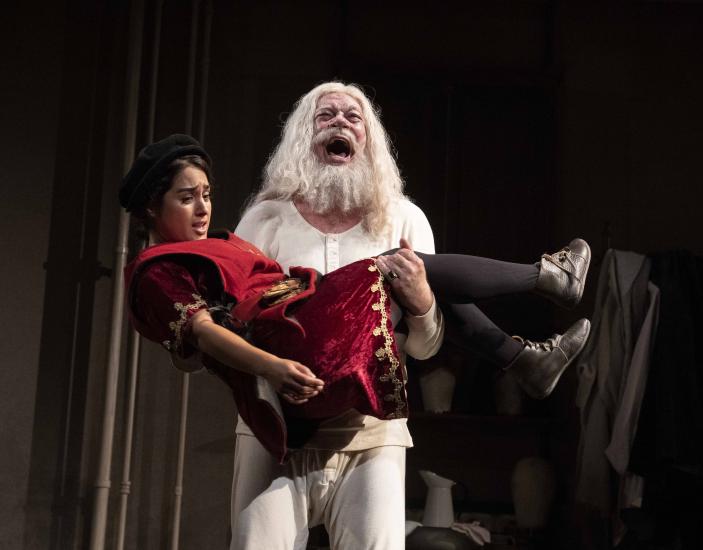 A man in white with white hair and a long white beard holds a jester in his arms, he is shouting
