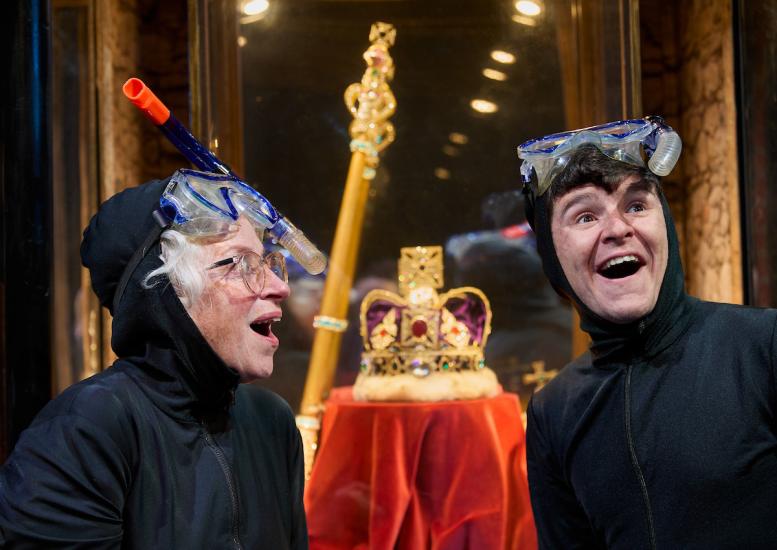 A Granny and a young boy wear balaclavas and stand in front of a glass cabinet with a crown inside