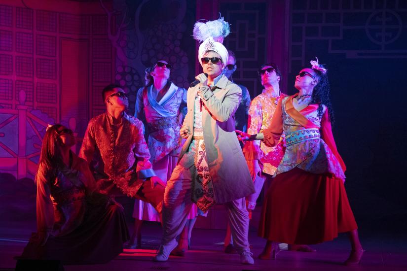 Aladdin is singing into a microphone with sunglasses on, the ensemble are also wearing sunglasses