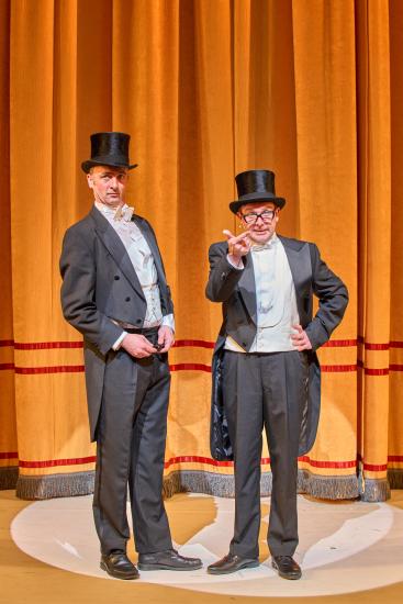 Two men in suits and top hats stand in front of an orange velvet curtain