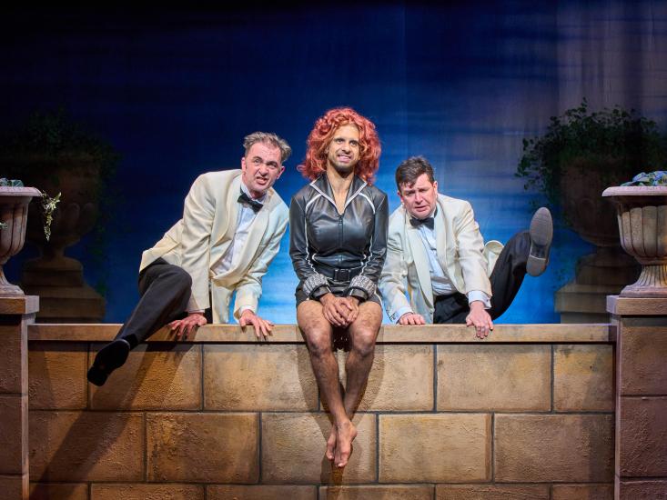 Two men in suits are trying to climb over a brick wall, another man in a tight leather dress and a red wig is sitting in between them