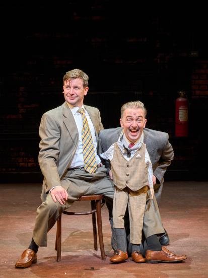 Two men in suits smile at the audience, one is sat on a chair and the other is on his knees with a small version of the suit tucked under his chin