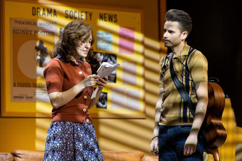 A woman and man stand in front of a Drama Society news board, the woman is reading from a book and the man has a guitar hanging on his back