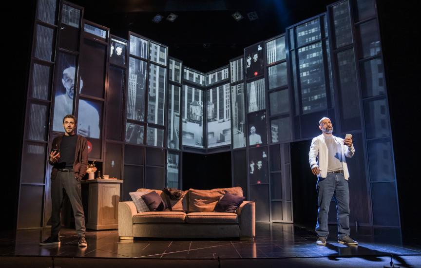 Two men stand in a living room either side of a sofa, they each have phones in their hands