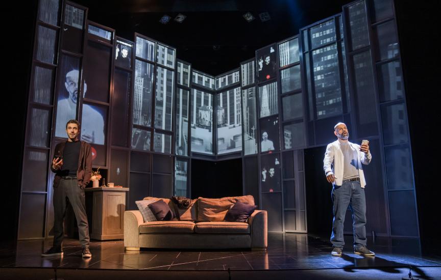 Two men stand in a living room either side of a sofa, they each have phones in their hands