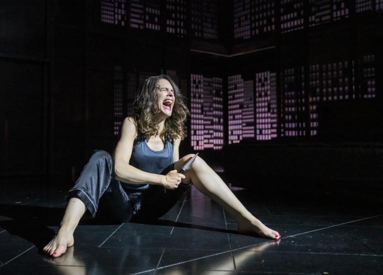 A woman sits on the ground screaming in pain, she holds a knife to her leg and blood is visible