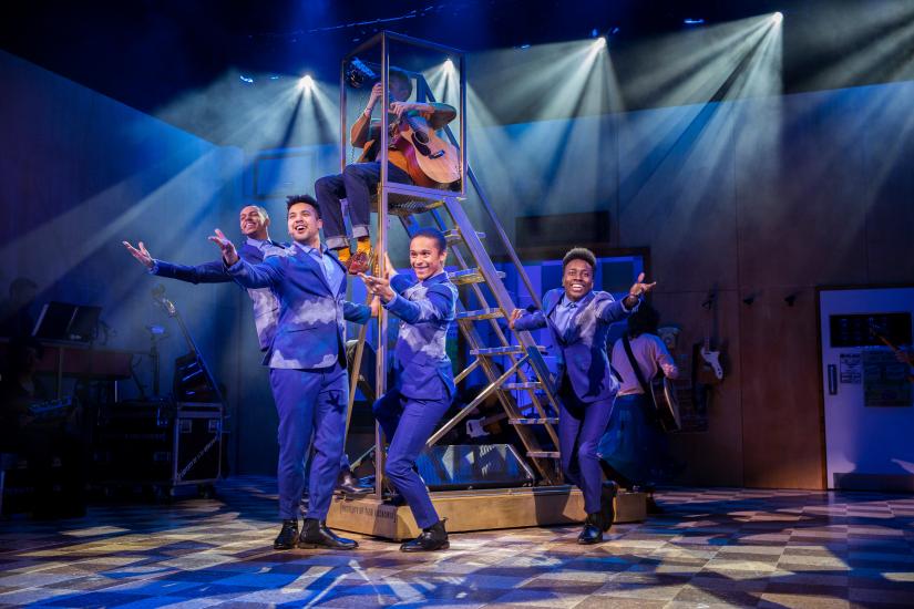 Four men in matching blue suits are singing and pointing to the audience, one is playing a guitar