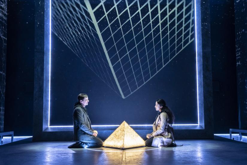 A man and a woman sit either side of a shining pyramid shape on the floor