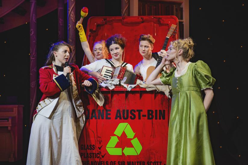 Five women in Regency dress are standing in a large bin, they are playing the accordion, guitar and a wooden maraca