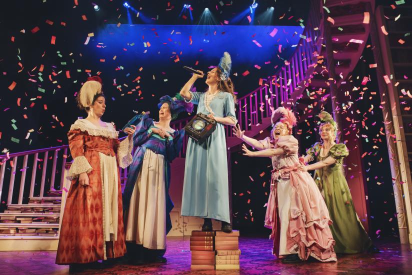 Four women in Regency dress are surrounding another, who is standing on a pile of books singing into a microphone. Confetti is falling around them 