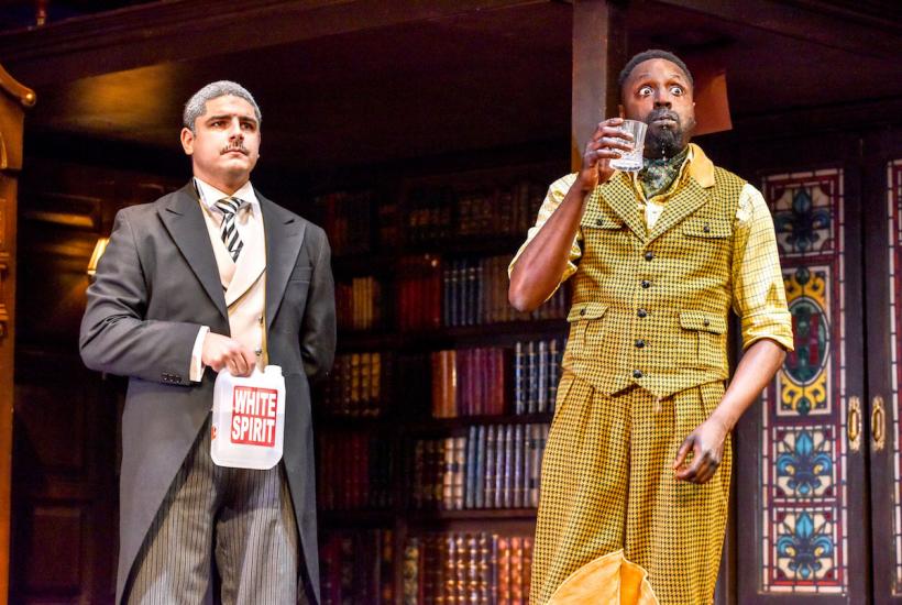 A man in a yellow dogtooth suit drinks from a tumbler looking shocked, a man next to him holds a bottle of white spirit