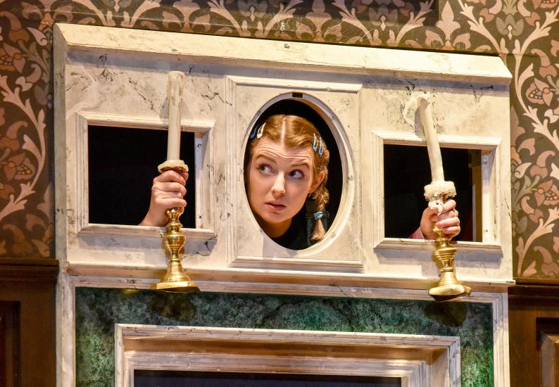 A woman holding two candlesticks, she has her face and arms through holes in a mantlepiece