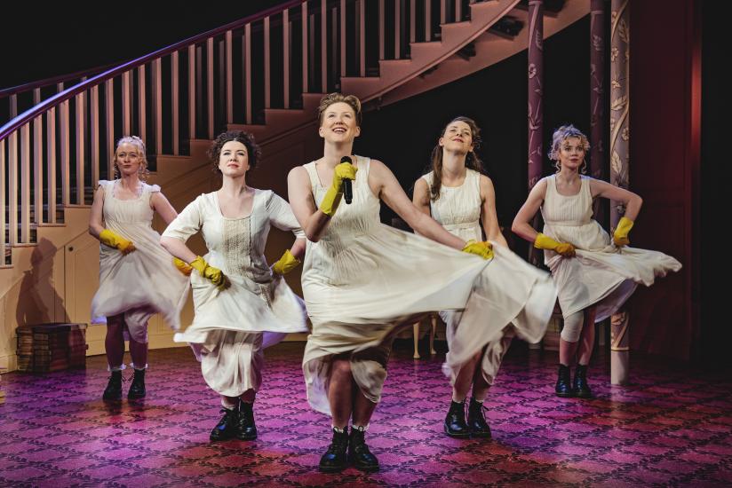 Five women in petticoats and yellow rubber gloves are singing and dancing in formation