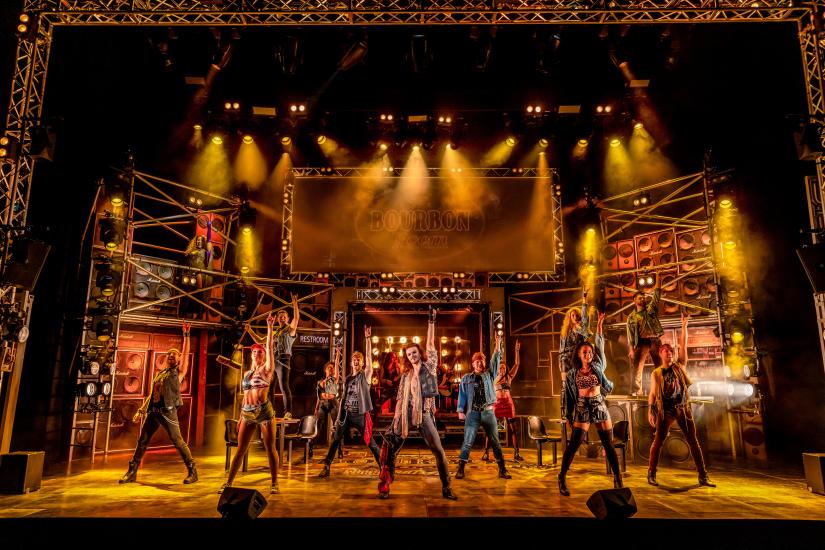 The cast of Rock of Ages dancing on stage, they are all doing the rock on hand sign