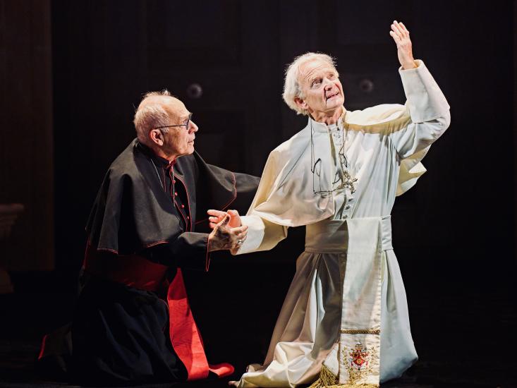 The two popes kneel together, one has his hand outstretched to the sky