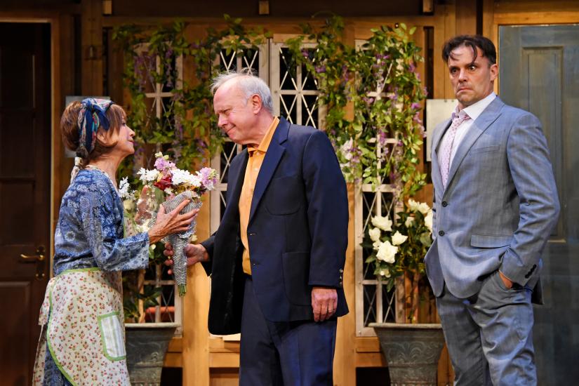 A man in a suit gives a bouquet of flowers to a woman, next to them a second man looks angry