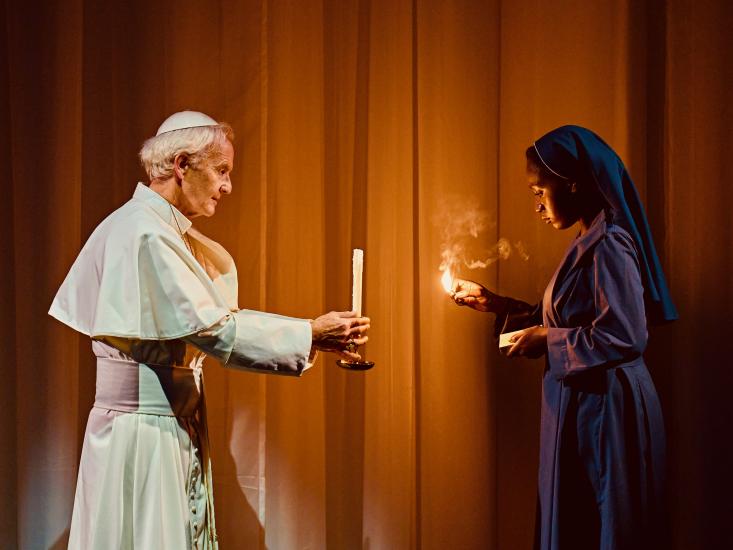 A pope in white dress holds a candle in front of a woman who holds a match