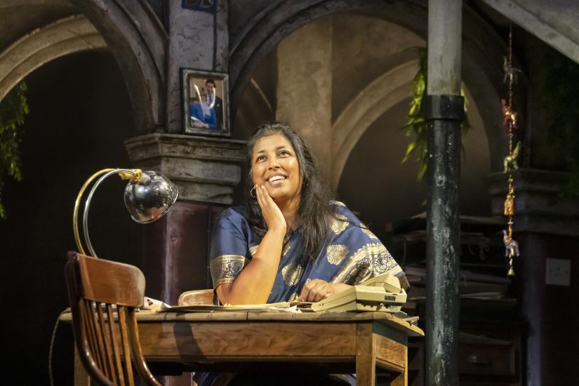 A woman wearing a sari sits at a desk, she is smiling with her head rested on her hand