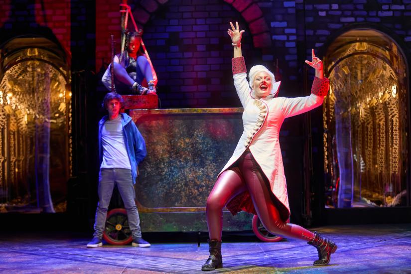 A white-haired woman dressed in red leggings and a white coat sings maniacally while an angry-looking girl is tied up in the background, an angry-looking boy standing by her.