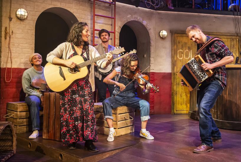 Five members of the company of Fisherman's Friends play instruments on stage, including a violin, a guitar, a banjo, and an accordion.