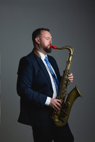 Dan Forshaw plays the saxophone in a navy suit and light blue tie.