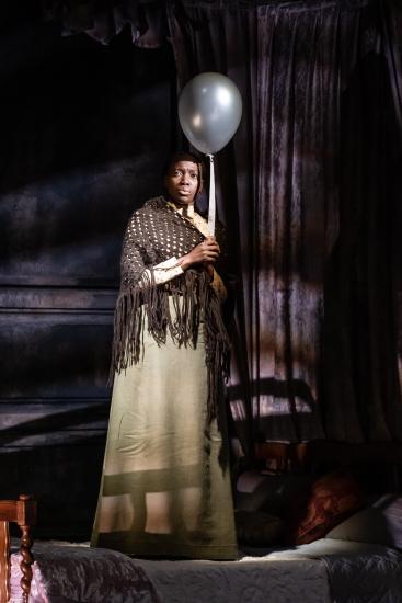 A woman in a long dress and brown knitted shawl holds a single silver balloon