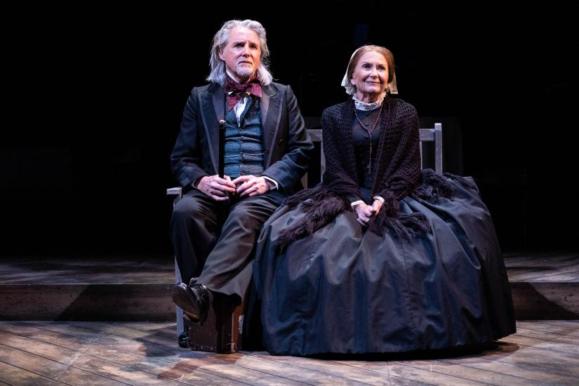 A man and woman in Victorian dress sit together on a wooden bench. The man has a suitcase by his feet.