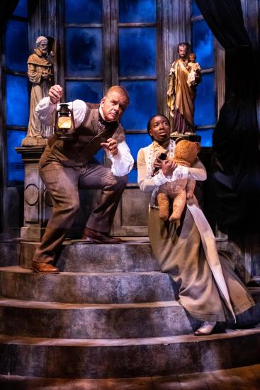 A man and woman in Victorian dress stand on a staircase. The woman holds a teddy bear and the man holds an oil lamp.