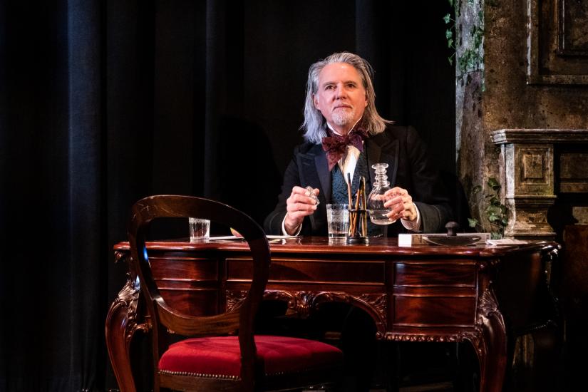 A man in Victorian dress sits at a writing desk with a decanter and whiskey glass