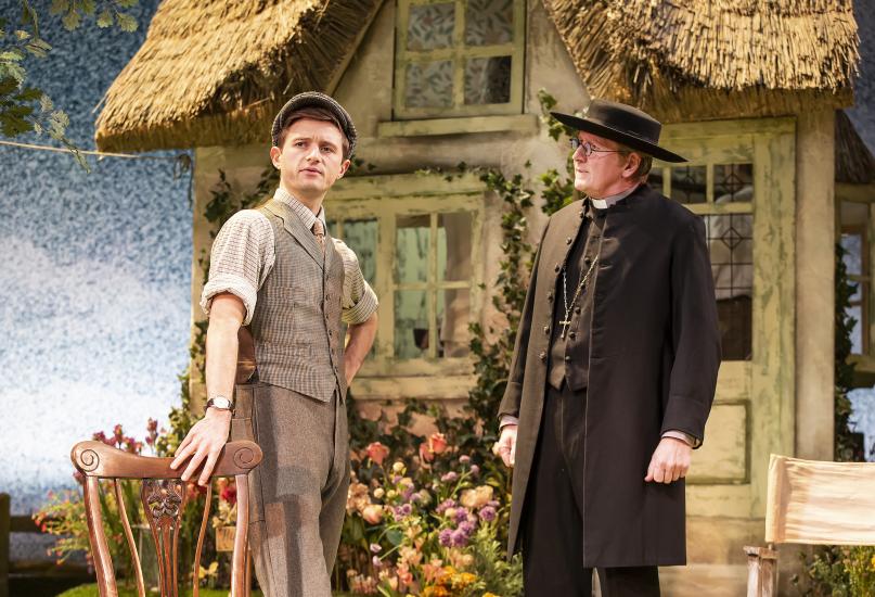 A smartly dressed gentleman talks with a reverend outside a thatched-roof house. 