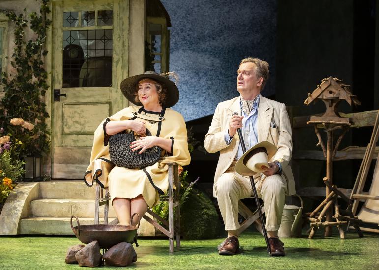 Mrs. Warren and Sir George Crofts sit in chairs outside a house in smart clothes. Mrs. Warren wears a large black hat. 