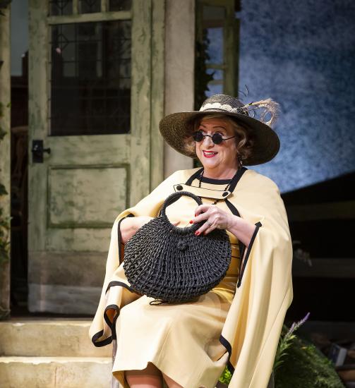 Mrs. Warren sits on a chair in a white dress with a black bag, sunglasses, and a large brimmed black hat. 