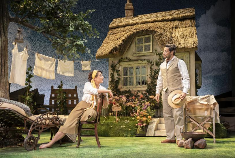 Vivie Warren lounges on a wooden chair, talking to Praed, a smartly dressed man in glasses. In the background there is a thatched-roof house, a washing line, and a garden. 