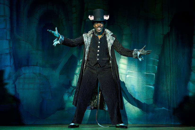 King Rat smiles with his arms wide, he is wearing a pinstripe suit and top hat