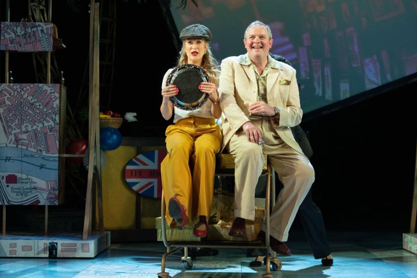 A woman in yellow trousers and a flat cap and a man in a light coloured suit suit on a table, while the woman holds a circular plate acting as a steering wheel