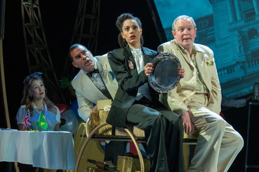 A woman in a black suit and a man in a light suit sit on a table pretending to be in a car. A man and a woman look on; all four have concerned expressions.