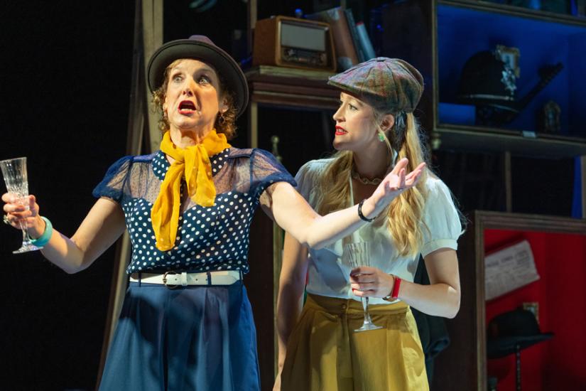 A woman in a spotty blue blouse and a yellow necktie holds a glass with her hands held out at either side, telling an extravagant story. Another woman in a flat cap stands at her side, listening. 