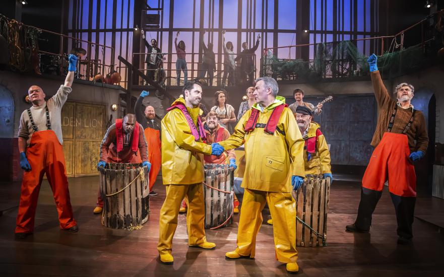 The company of Fisherman's Friends cheer on stage as two men in yellow raincoats shake hands.